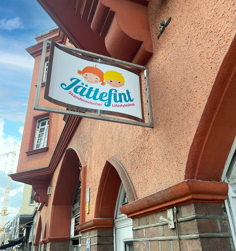 Schild des skandinavischen Lifestyleshops Jättetint, das fröhliche Kinderillustrationen zeigt, vor einer farbenfrohen Wand. Ideal für skandinavische Wohnaccessoires und Kinderprodukte. Corporate Design Köln