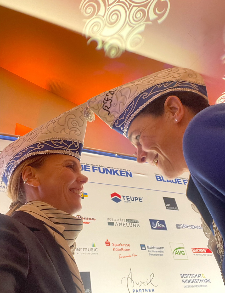 Zwei Frauen in traditionellen Kostümen mit Hüten schauen sich lächelnd an. Im Hintergrund sind Sponsorenlogos einer Veranstaltung sichtbar, die die "Blauen Funken" repräsentiert.