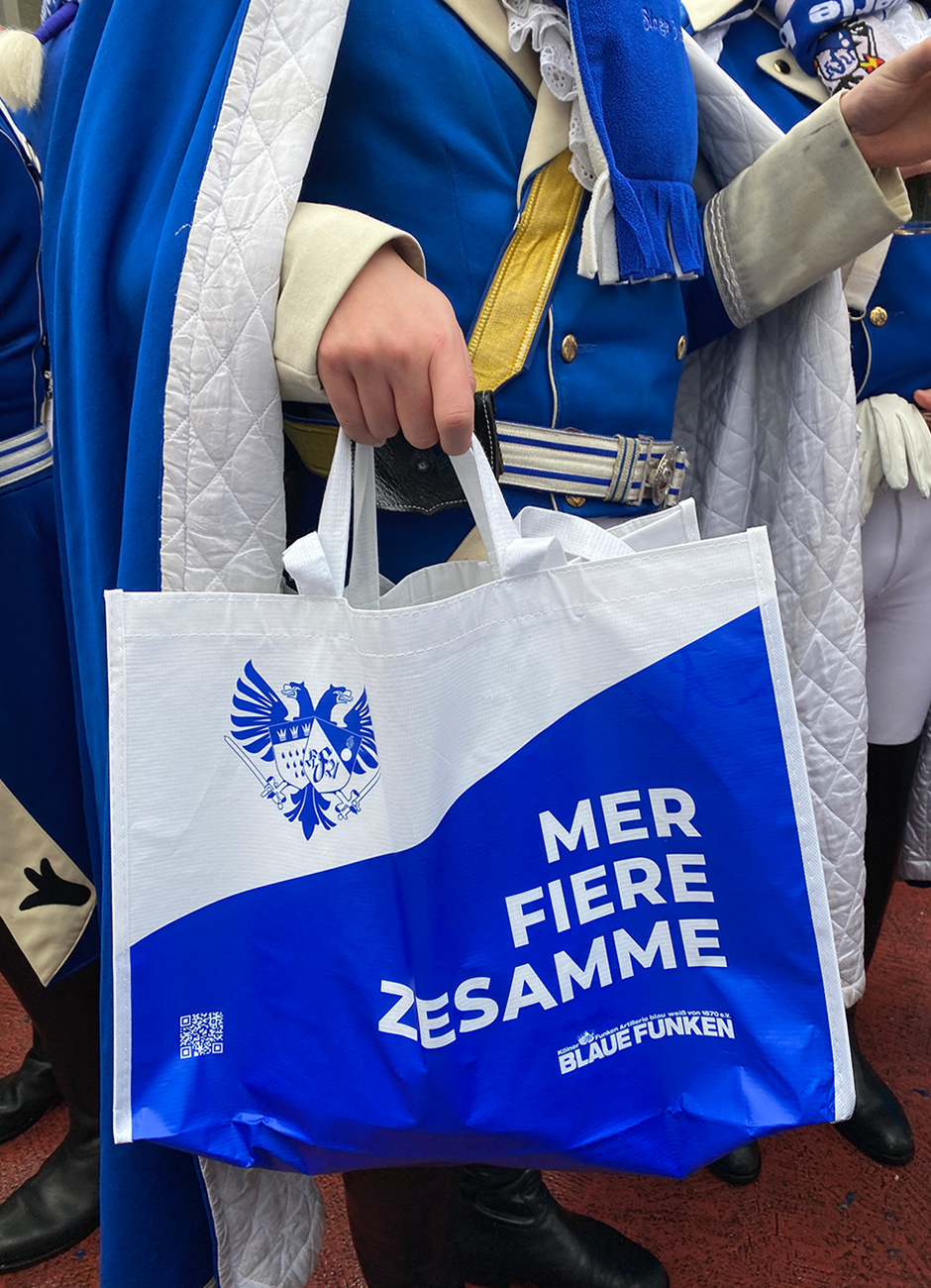 Eine Person hält eine weiße Tragetasche mit blauem Aufdruck, auf dem steht "MER FIER ZESAMME", während sie in traditioneller Kleidung der Blauen Funken auf einem Fest steht.