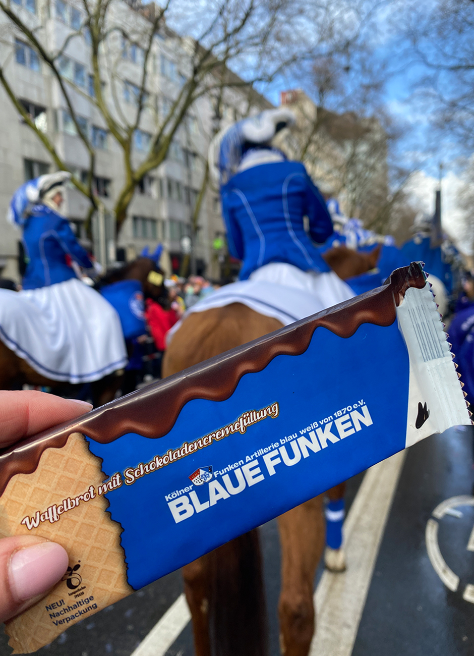 Verpackung der Waffelschnitte "Blaue Funken" mit Schokoladencremefüllung, gehalten vor einer Karnevalsparade mit Reiterinnen in blauen Kostümen.