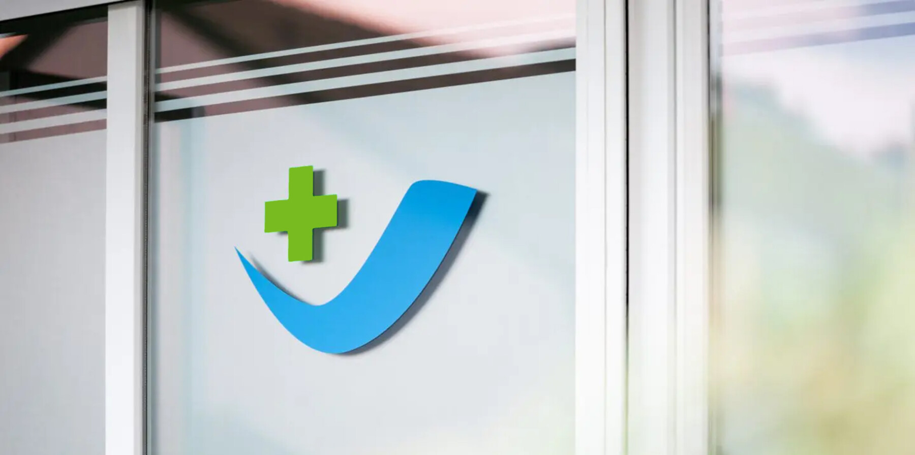Logo einer Arztpraxis mit einem grünen Kreuz und einer blauen Wellenlinie, das an einer Glaswand angebracht ist. Symbolisiert medizinische Dienstleistungen und Gesundheitsversorgung.