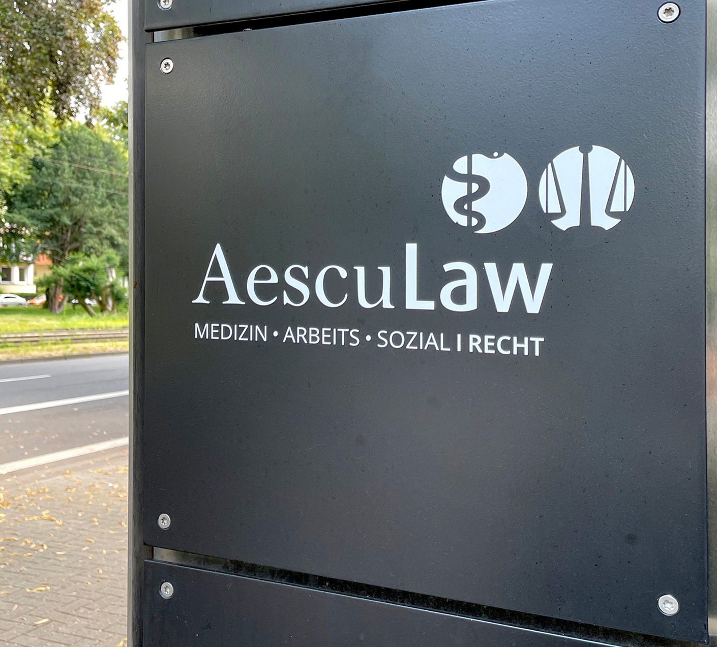AescuLaw Schild mit den Schwerpunkten Medizin, Arbeitsrecht und Sozialrecht, präsentiert auf modernem Hintergrund in städtischer Umgebung. Rechtsberatung für individuelle Anliegen.