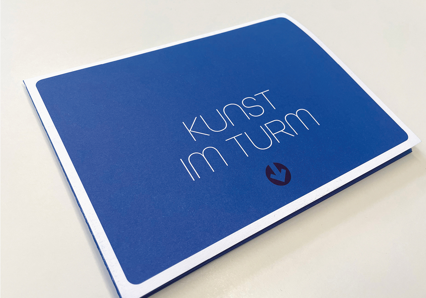 Blaue Broschüre mit dem Titel Kunst im Turm, die Informationen zu einer Kunstausstellung oder Veranstaltung enthält. Modernes Design mit klarer Schrift.