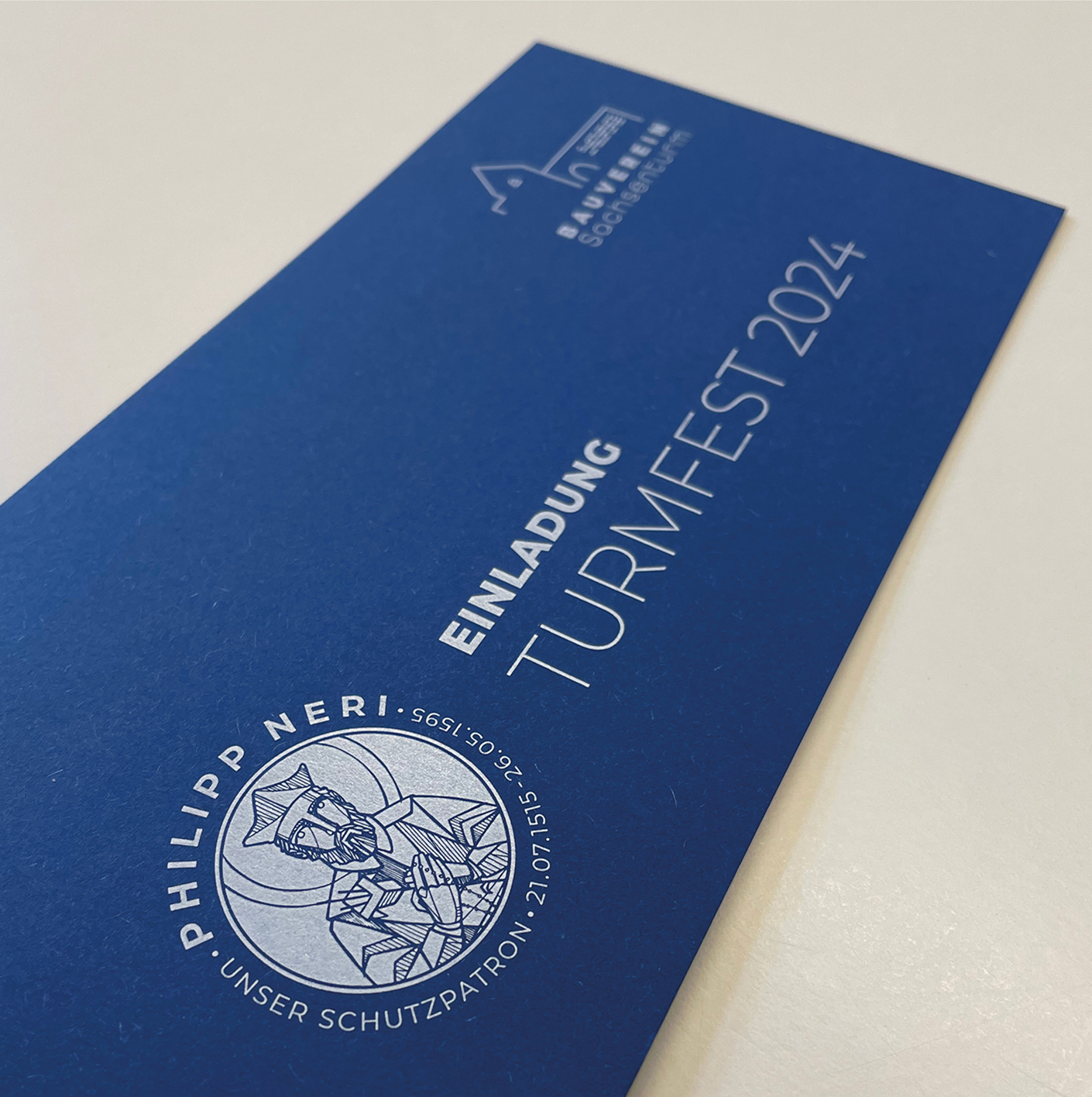 Einladungskarte zum Turmfest 2024 mit dem Logo von Philipp Neri, dem Schutzpatron. Das Design ist in Blau gehalten und vermittelt festliche Vorfreude auf die Veranstaltung.