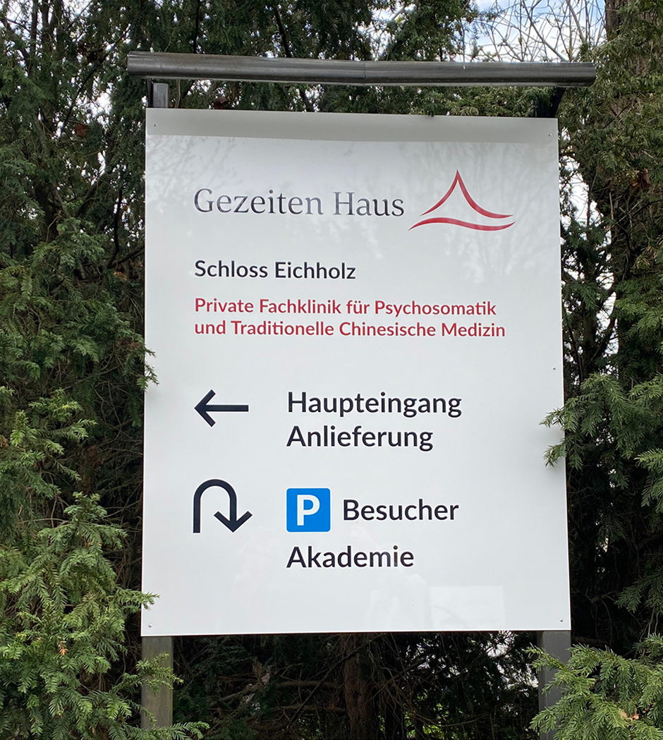 Hinweisschild des Gezeiten Hauses, einer privaten Fachklinik für Psychosomatik und Traditionelle Chinesische Medizin in Schloss Eichholz, mit Richtungsanzeigen für Haupt- und Besucherzugang.