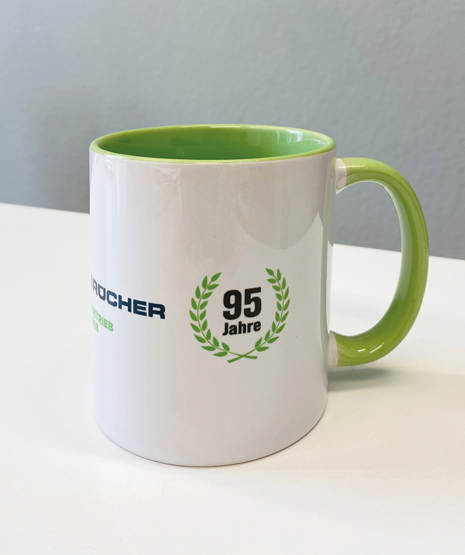 Tasse mit grünem Innenraum und Henkel, bedruckt mit dem Jubiläumslogo "95 Jahre" und dem Namen des Unternehmens. Ideal für Kaffeegenuss im Büro oder zu Hause.