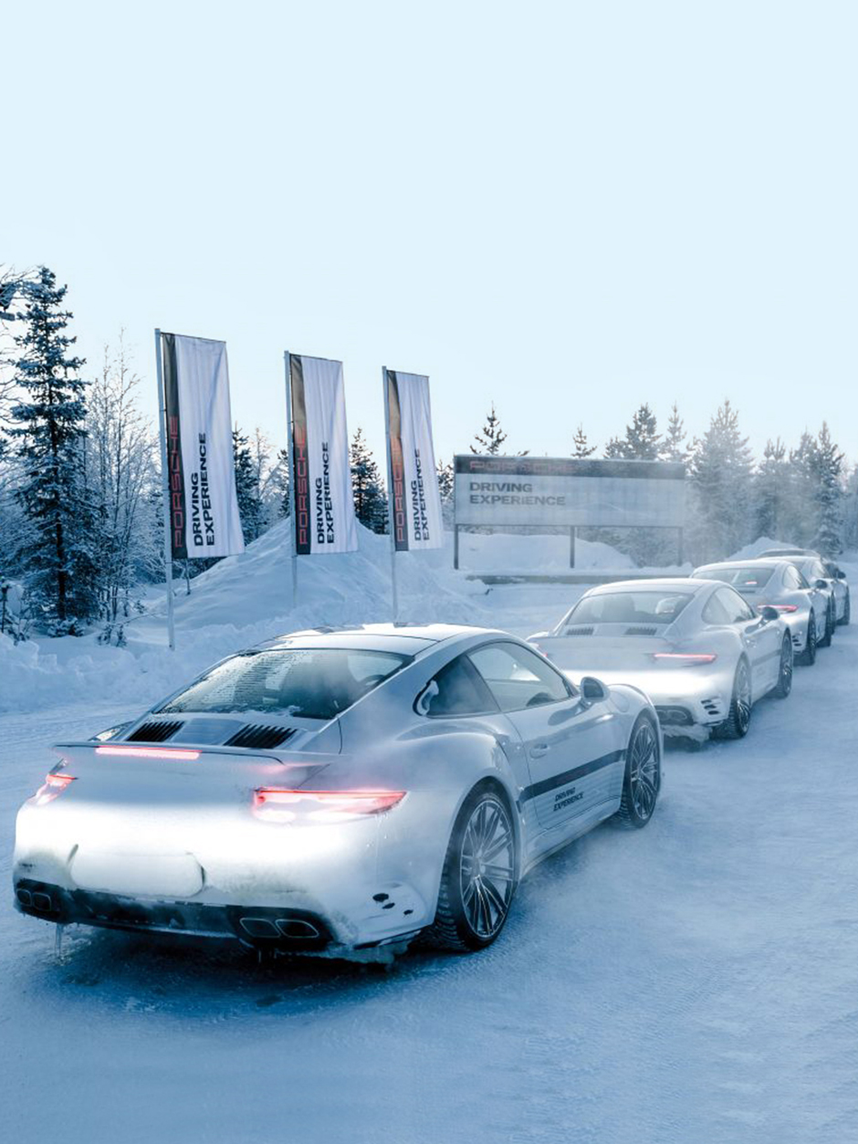 Porsche Driving Experience im Schnee mit mehreren Fahrzeugen. Die Autos stehen in einer Reihe auf der verschneiten Straße, umgeben von winterlicher Landschaft und Werbebannern.