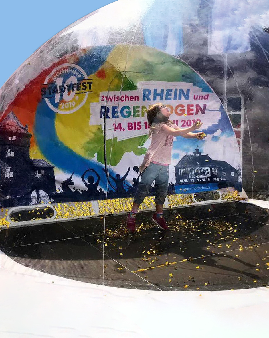 Kind in einer transparenten Kugel beim Stadtfest Monheim 2019. Hintergrund zeigt das Event-Logo sowie bunte Grafiken. Stimmungsvoll und fröhlich, umgeben von bunten Blütenblättern.