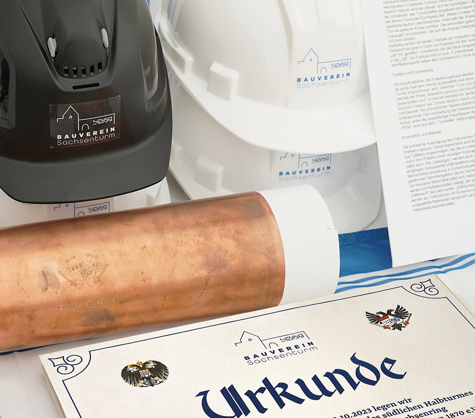 Bauhelm, Urkunde und Bauunterlagen des Bauvereins Sachsenzentrum auf einem Tisch, symbolisieren die Feier eines Bauprojekts und die Bedeutung des Bauens in der Region.