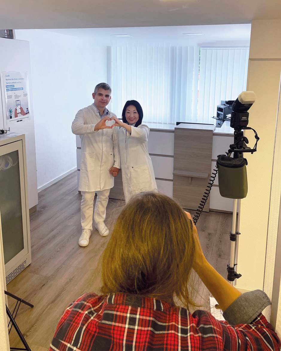 Zwei Ärzte in weißen Kitteln bilden ein Herz mit ihren Händen. Im Hintergrund befindet sich eine moderne Praxis. Eine Fotografin nimmt das Bild auf.