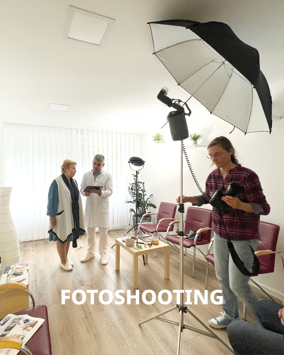 Fotoshooting in einem modernen Raum mit medizinischer Atmosphäre. Ein Arzt bespricht etwas mit einer älteren Dame, während eine Fotografin die Szene festhält. Beleuchtung und Requisiten sind sichtbar.